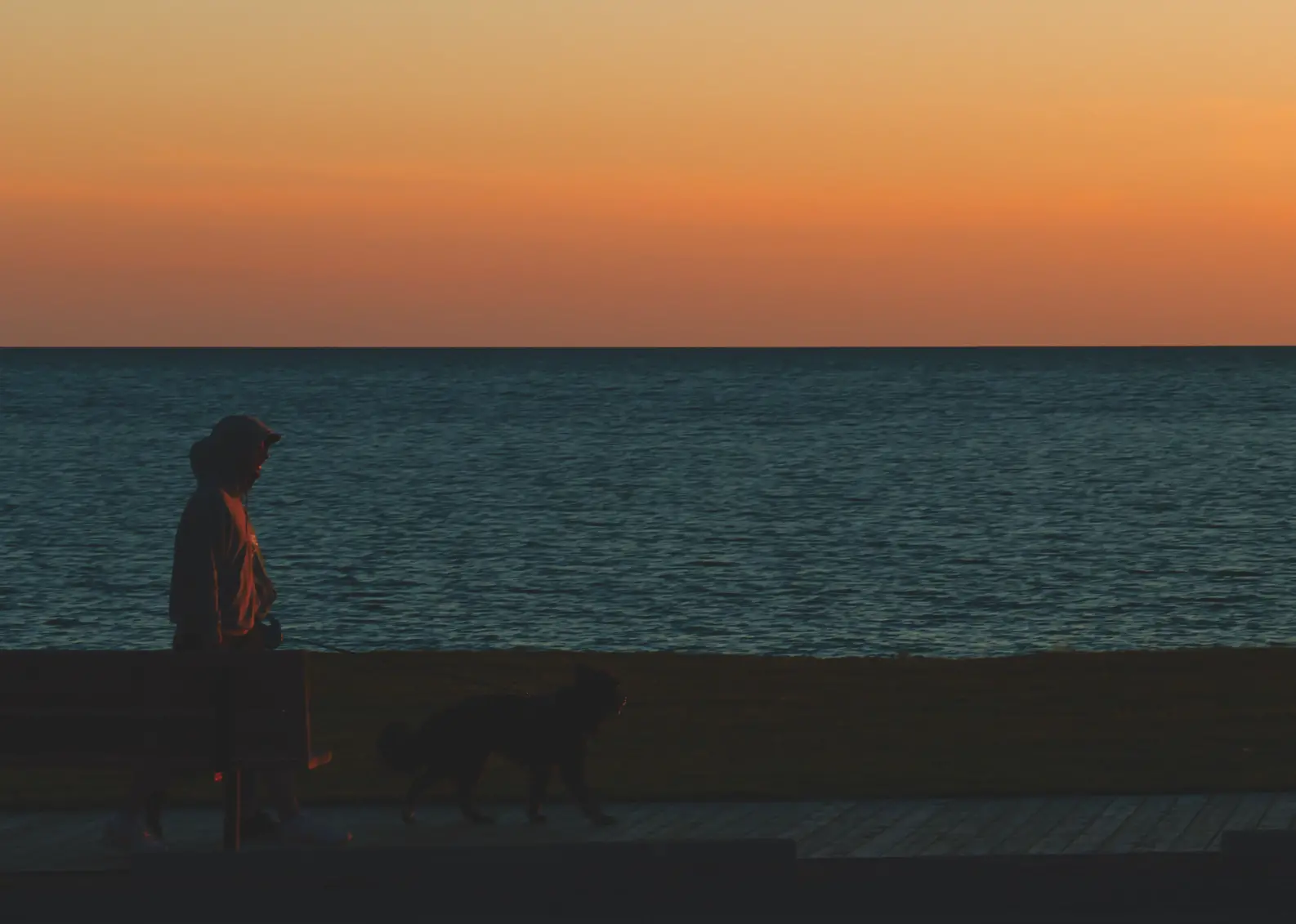 A couple walk their dog along the boardwalk next to the sea under a vibrant orange sunset.