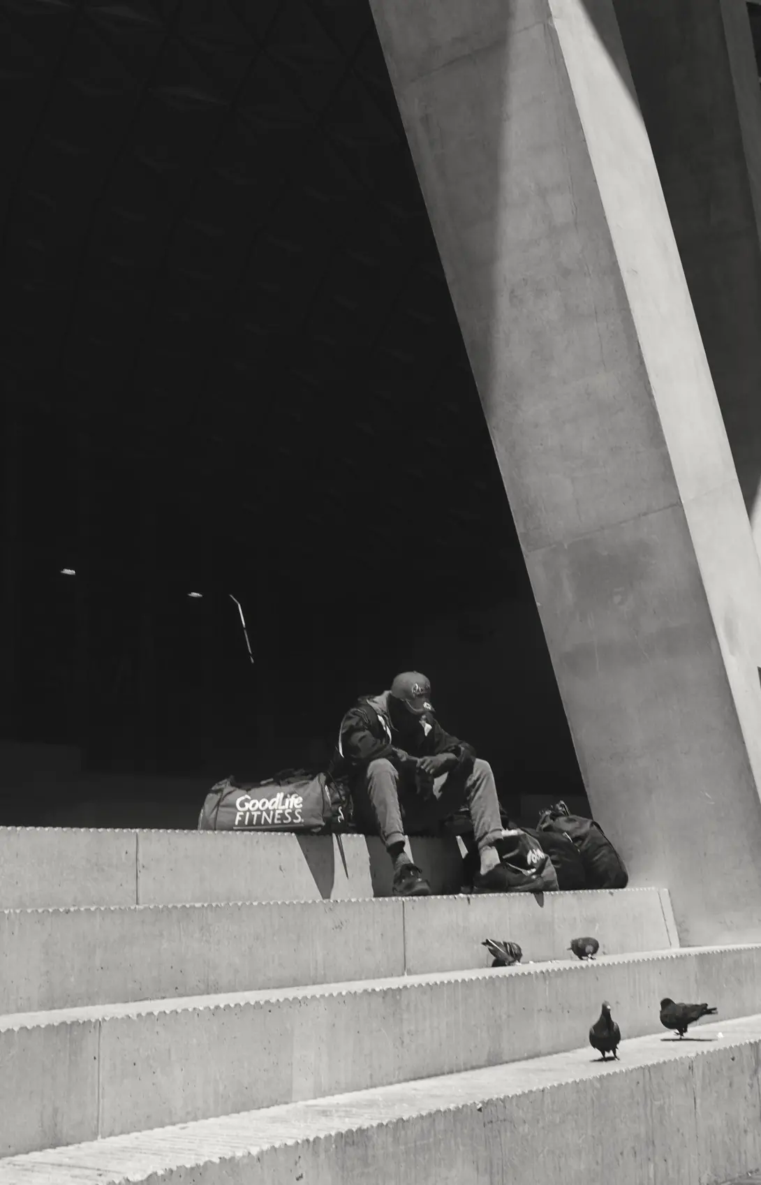 Black and white photo of a lone man sitting on the steps of the Toronto Metropolitan University watching a group of pigeons in front of him. The bag next to him says Good Life.