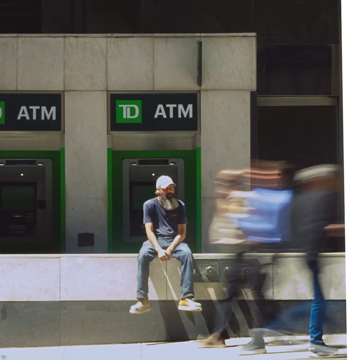 A man with a bushy beard sits in front of an ATM on the stree watching people pass by.