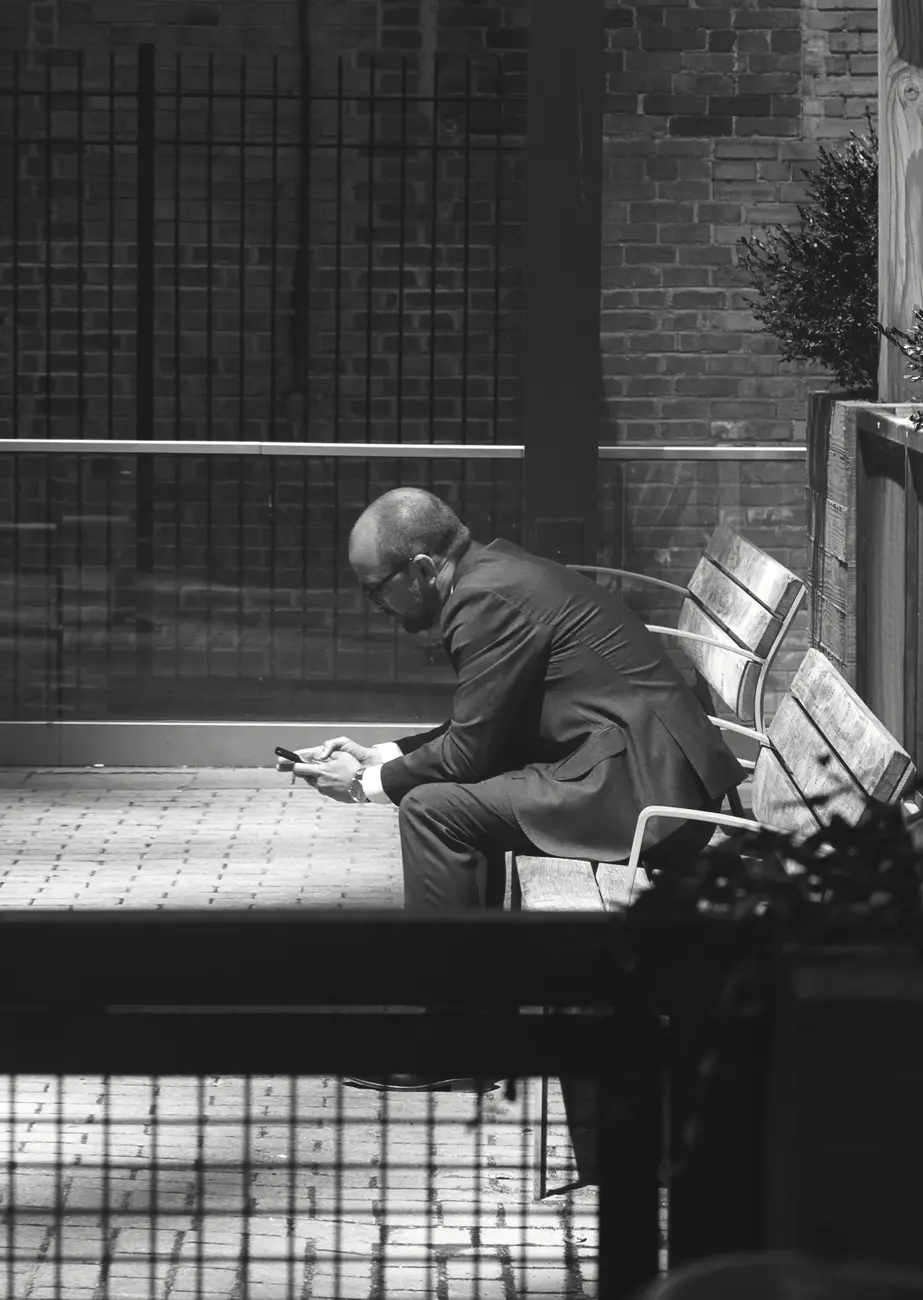 Black and white shot of a man sitting on a bench looking at his phone while on smoke break.