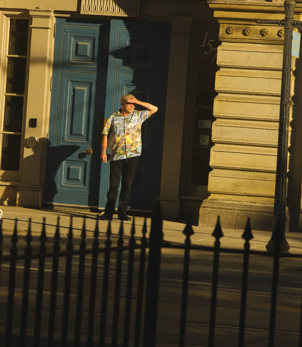 A man in a tropical shirt stands outside of a building with two blue doors shielding his eyes from the rays of the sun.