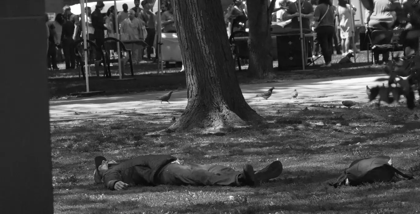 A man fully asleep in the park with mouth agape as if he had just died there. A large crowd can be seen behind him attending a small festival.