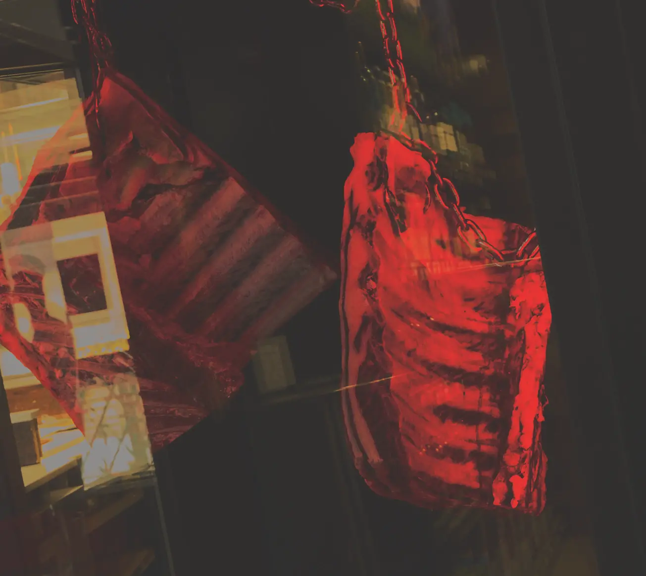 Two racks of ribs hang from chains in a freezer with a black background and dim red light shined on them.