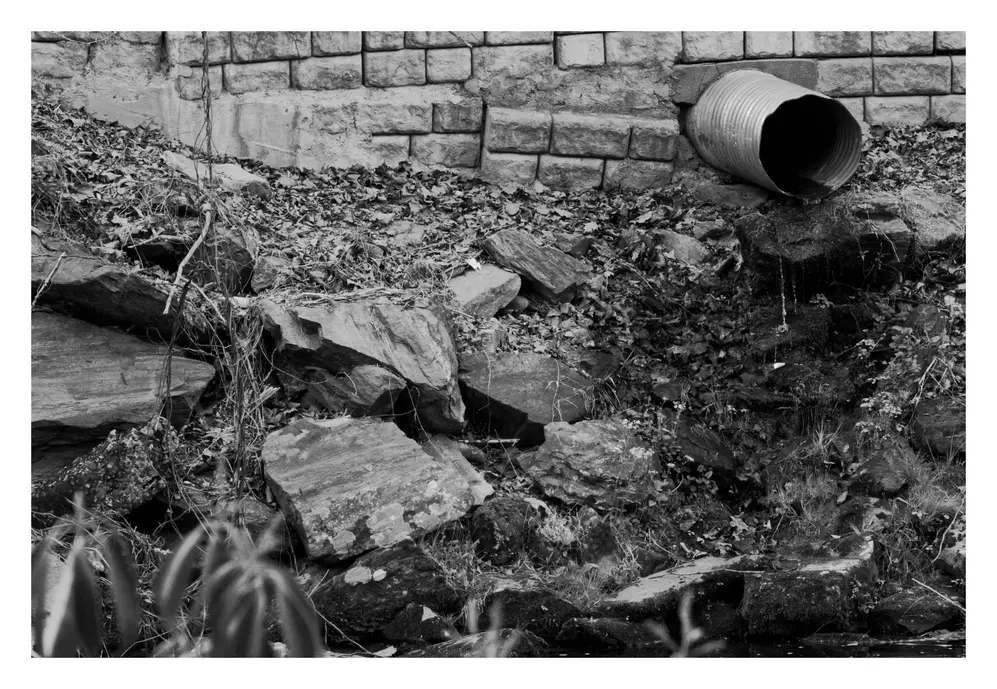 black & white photo of a drainage pipe going into a nearby river. The water trickles down the various rocks below.